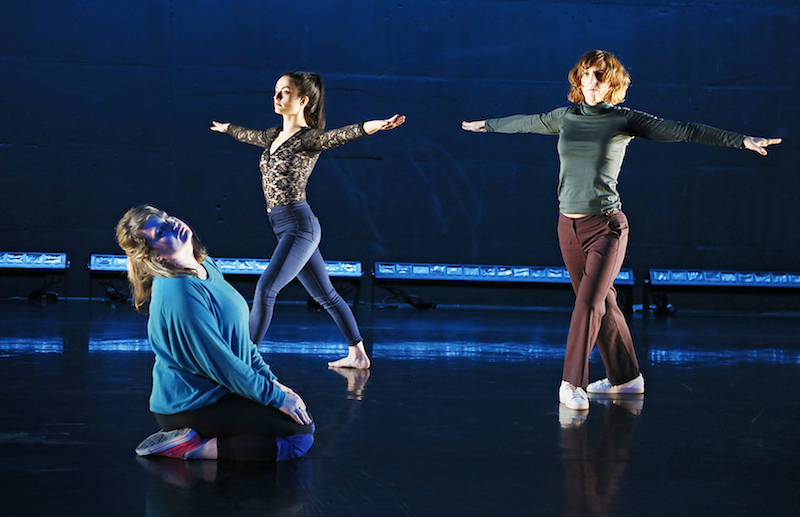 Two performers in pants and long sleeve shirts stand in a lunge with their arms stretching out at their sides. A third performer sits on her shins and her head extends back.